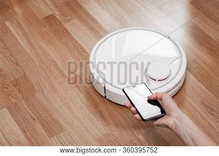 Housewife Using Mobile To Control Robotic Vacuum Cleaner. Robot Controlled By Voice Commands To Dire