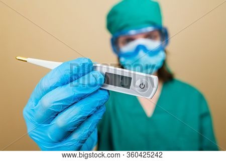 Female Doctor In Green Suit And Blue Mask Gloves And Protective Glasses Withthermometer In Her Hand 