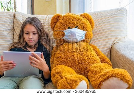 Beautiful Little Girl In A Black Jacket Playing With Her Brown Teddy Bear Who Has Put A Mask To Prot