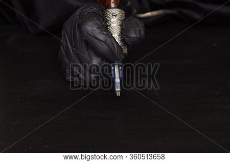 Part Of A Rotary Tattoo Machine With A Needle In Hand In A Black Glove On A Black Background. Side V