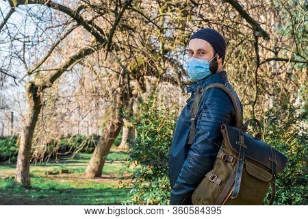 Adult Bearded Man With Medical Mask On His Face Walks Through Park During The Pandemic.