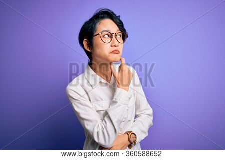 Young beautiful asian girl wearing casual shirt and glasses standing over purple background Thinking worried about a question, concerned and nervous with hand on chin