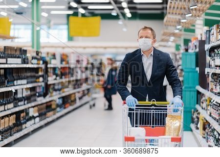 Photo Of Adult Man Wears Disposable Face Mask, Makes Shopping, Thinks About Protection And Prevent M