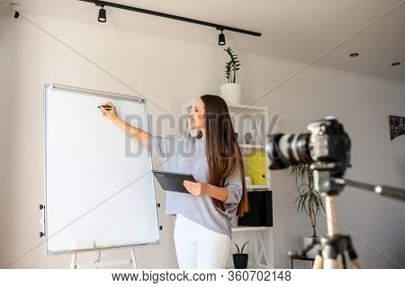 Young Woman Teacher In Casual Wear Makes Tutorial Video On The Camera On A Tripod Indoors . She With