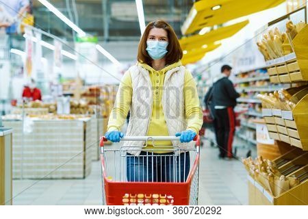 Girl In Medical Mask And Gloves Standing With Shopping Cart Between Aisles At Grocery Store And Look