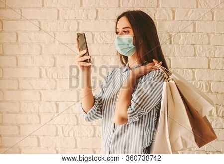Beautiful Young Woman Shopaholic In Medical Face Mask Hold Mobile Phone And Shopping Bags In Hands. 