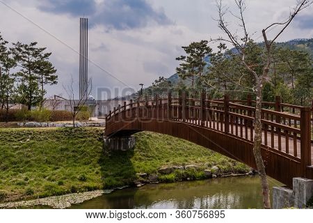 Hwanggan, South Korea; April 10, 2020: Side View Of Wooden Footbridge Over Small Stream With War Mem