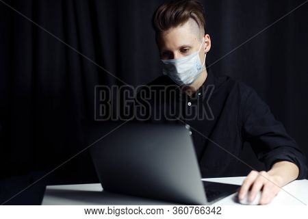 A Young Freelancer Works From Home. The Guy Is Sitting At Home In A Protective Mask, Self-insulating