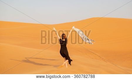 A Girl Walks On The Sand In A Deserted Desert. A Woman In A Black Dress With A Cloth In Her Hands In