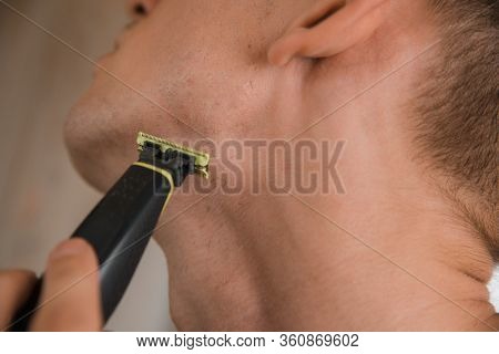 Close Up Of Young Man Looking To Mirror And Shaving Beard With Trimmer Or Electric Shaver
