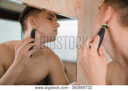 Close Up Of Young Man Looking To Mirror And Shaving Beard With Trimmer Or Electric Shaver