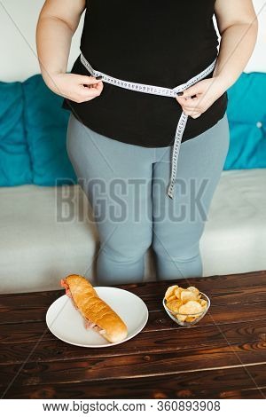 Imbalanced Nutrition, Weight Gain, Dieting. Overweight Woman With Sandwich, Chips And Measuring Tape