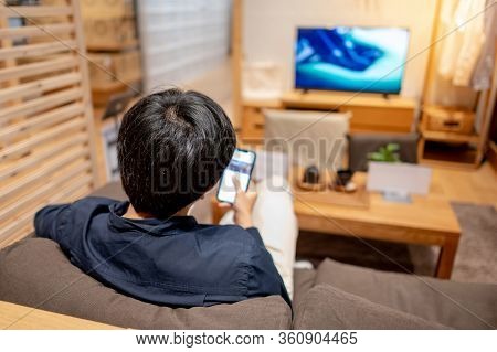 Young Asian Man In Casual Clothing Sitting On Comfortable Sofa (couch) Using Smartphone And Watching