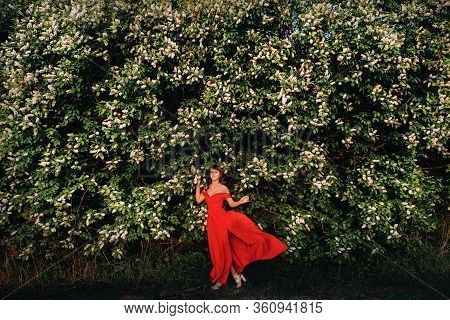 A Beautiful Girl In Spring In A Red Dress With Red Lips Stands Next To A Large White Flowering Tree.