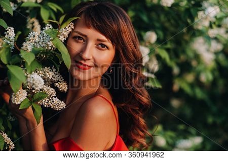 A Beautiful Girl In Spring In A Red Dress With Red Lips Stands Next To A Large White Flowering Tree.