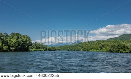 A Calm Blue River Between The Banks Overgrown With Lush Green Vegetation. A Mountain Range Against A