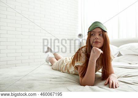 Young Focused Caucasian Girl Lying And Looking Away On Bed At Home In Morning Time. Beautiful Red Ha
