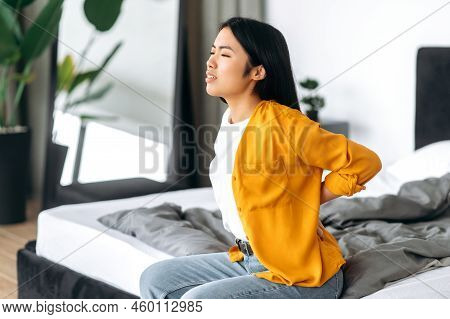Back Pain, Back Spasm. Young Asian Brunette Woman In Stylish Clothes, Sits On A Bed In A Room, Massa