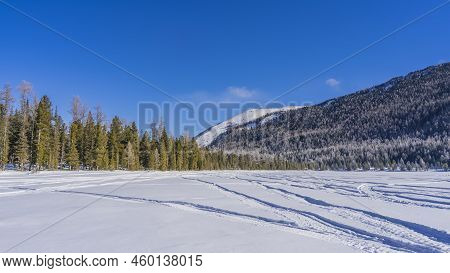 In A Snow-covered Valley, On The Surface Of A Frozen Lake, Snowmobile Tracks Are Visible. Coniferous