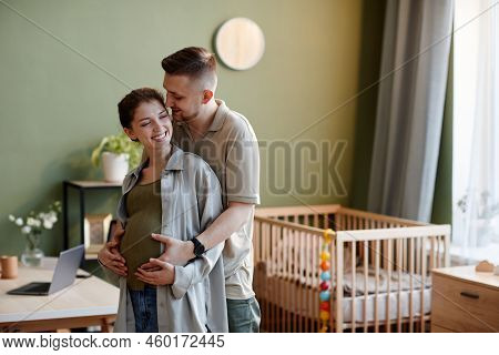 Young Man Embracing His Pregnant Wife Hile They Standing In Kids Room At Home