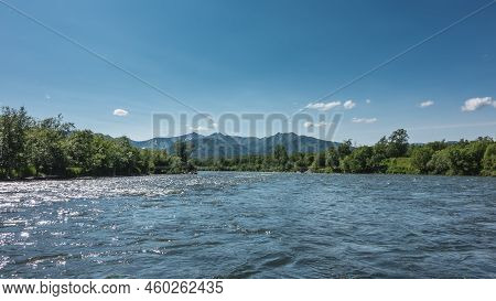 The Blue River Sparkles In The Sun. There Is Lush Green Vegetation On The Banks. A Mountain Range Ag