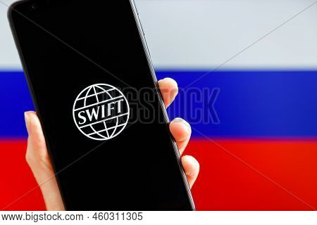 Ukraine, Kiev - February, 23 2022: Hand Holding Mobile With Swift Logo And Russian Flag In Backgroun