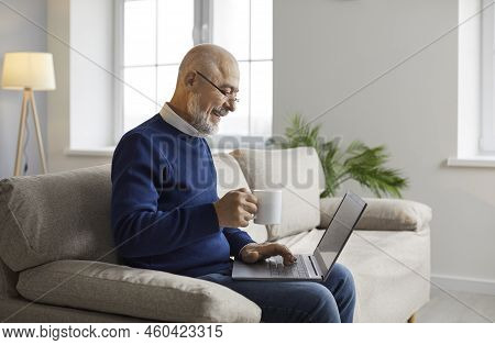Happy Senior Man Sitting On Sofa At Home, Using Laptop Computer And Drinking Coffee