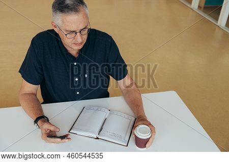 High Angle View Of Mature Male Entrepreneur Using Smartphone And Holding Coffee Cup By Diary On Desk