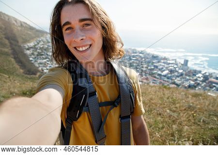 Portrait Of Smiling Young Male Explorer With Backpack Standing On Mountain With Cityscape, Sea And C