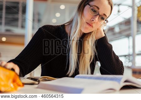 Focused Young Caucasian Woman With Blond Hair Wearing Eyeglasses And Studying At Desk In Library At 
