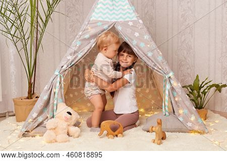 Portrait Of Cute Little Sisters Playing In Wigwam Hugging While Playing Together, Elder Girl And Tod