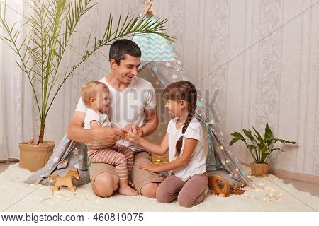 Indoor Shot Of Smiling Brunette Man Wearing White T Shirt Sitting In Wigwam With His Daughters And H