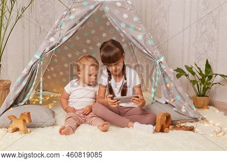 Image Of Calm Little Sisters Wearing White T Shirts Sitting In Wigwam And Watching Cartoons Together