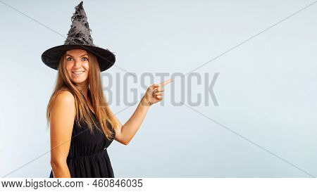 Woman In Witch Costume Pointing With Finger At Copyspace Side And Looking At Camera On Blue Backgrou