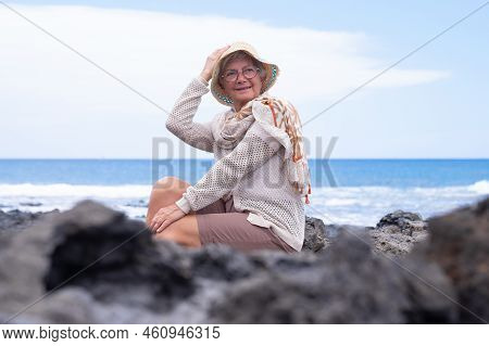 Smiling Caucasian Senior Woman With Hat Sitting At The Beach Looking Away Enjoying Vacation Or Retir