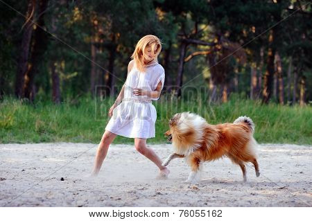 Beautiful girl with blonde hair in a white dress running on the sand and playing with her fluffy col