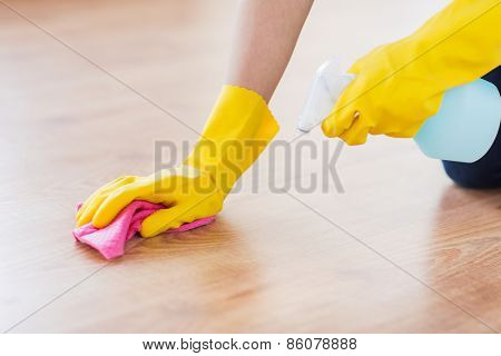 people, housework and housekeeping concept - close up of woman in rubber glover with cloth and detergent spray cleaning floor at home