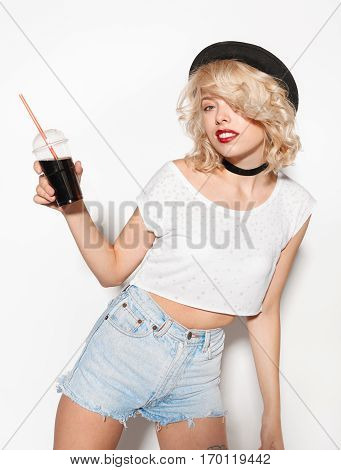 Fashion girl holding a glass with soda. Stylish blond with red lips and summer clothes drinking sweet soda. Attractive young woman in hipster outfit