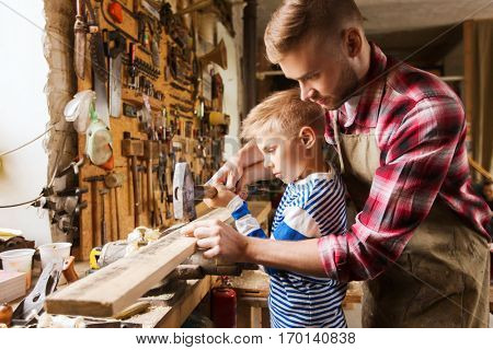 family, carpentry, woodwork and people concept - father and little son with hammer hammering nail into wood plank at workshop