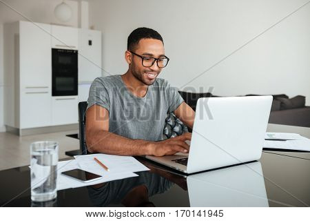 Photo of happy african man dressed in gey t-shirt and wearing eyeglasses using laptop and sitting at the table.