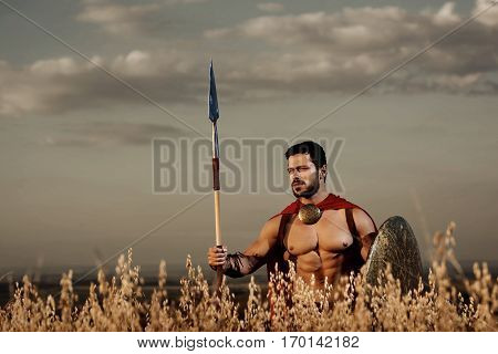 Athletic warrior with beard wearing like spartan among grass in field, holding iron weapon. Brunet soldier with bare torso in red cloack going in attack with rounded shield and weapon. Gray sky.