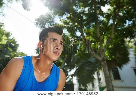 Low angle view of Vietnamese sportsman in headphones taking break after energy-sapping training in sunny park