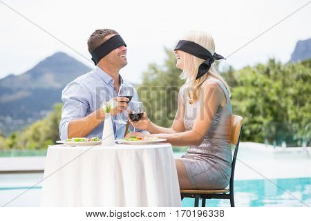 Blindfolded couple holding red wine while sitting by swimming pool at resort