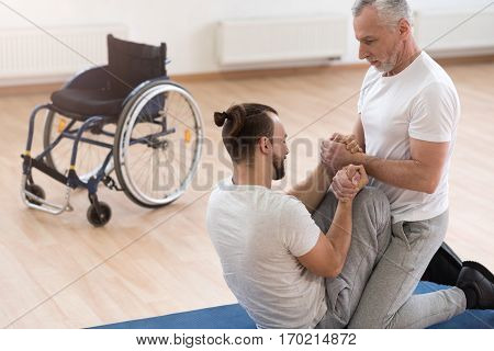 Developing strength. Openhearted athletic powerful orthopedist stretching the handicapped and assisting him while holding arms of the patient