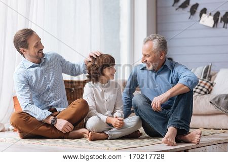 My boy. Positive nice delighted man laughing and pulling his sons hair while sitting on the floor with his family