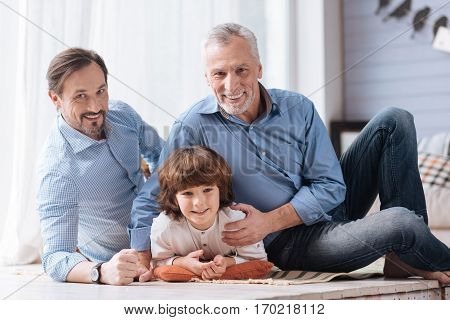 Feeling comfortable. Nice cute positive child lying on the cushion and smiling while being hugged by his grandfather