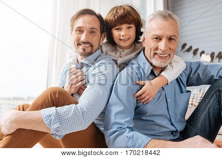Family bounds. Nice delighted cute boy hugging his dad and grandfather and looking at you while standing behind them
