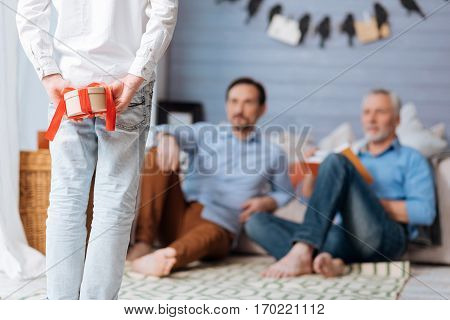 Preparing a surprise. Nice young pleasant boy standing and looking at his father and grandfather while holding a present behind his back
