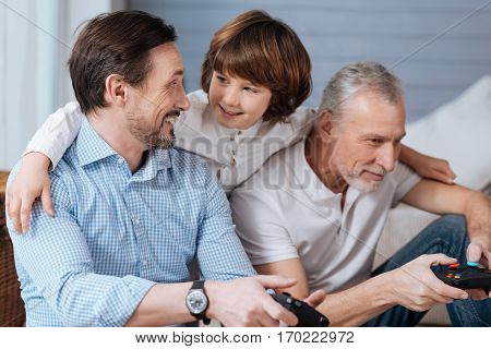 Great time together. Cute positive young boy standing behind has father and grandfather and hugging them while watching their play