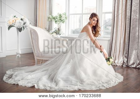 Wedding. Bride in beautiful dress sitting on sofa indoors in white studio interior like at home. Trendy wedding style shot in full length. Young attractive caucasian brunette model like a bride against big window tender posing.
